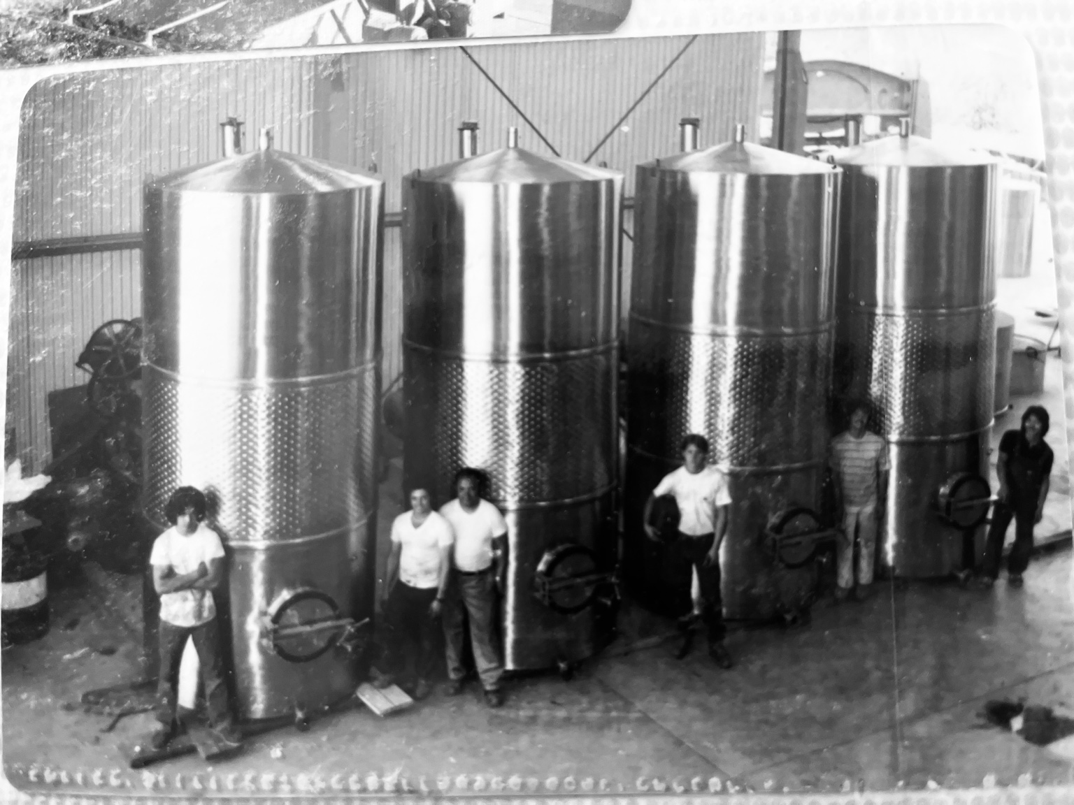 Historical photograph of four stainless-steel winery tanks and workers